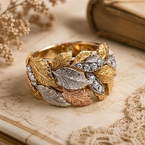 May include: A wide band ring featuring a textured leaf motif crafted in yellow, white, and rose gold tones. The design incorporates multiple overlapping metal leaves with a detailed vein pattern, accented by small, round, clear, sparkling gemstones.