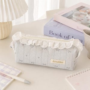 Op de afbeelding: Een rechthoekig etui van lichtgrijze stof met een herhalend patroon van kleine strikjes en verticale stippellijnen. De bovenrand is versierd met een witte kanten ruche en een ritssluiting. Een klein rechthoekig label bevat de tekst Sezakka.