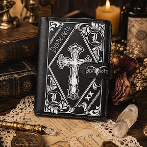 May include: A black rectangular notebook featuring a white printed design of a central cross, ornate corner flourishes, the letter L in circles, and the text Death Note. The cover includes a magnetic strap closure also labeled with Death Note.