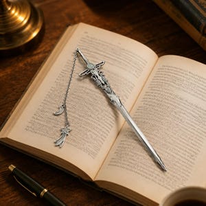 May include: A silver-toned metal bookmark shaped like a sword with an ornate hilt featuring a small circular inlay. A delicate chain is attached to the hilt, dangling a crescent moon charm and a shooting star charm with a textured surface finish.