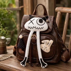 Puede incluir: Una mochila de piel sintética marrón oscuro con solapa y cierre de hebilla. En la parte frontal tiene un personaje de peluche blanco con un ojo en forma de X, un ojo de botón y brazos largos colgantes, junto a un conejo de tela y un dije.