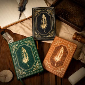 May include: Three hardback journals in black, green, and brown, each featuring a gold-foiled feather illustration on the front cover, surrounded by an ornate gold-foiled border and a textured background pattern on the front and back covers.
