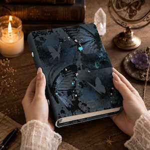 May include: A rectangular notebook featuring a dark blue and black cover with a printed butterfly pattern, paint splatter details, and light blue accents. The notebook has a smooth finish and a secure side closure mechanism with a visible white button.