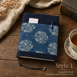 May include: A navy blue fabric book cover featuring a central band with a light blue floral medallion pattern. The cover includes a small pocket containing a white card, a blue pen holder, and a dark blue cord with two silver beads at the bottom.