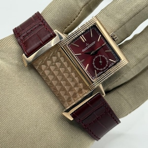 Op de afbeelding: Een rechthoekig goudkleurig horloge met een dieprode wijzerplaat voorzien van het Jaeger-LeCoultre logo en een kleine secondewijzer. Het horloge heeft een getextureerde gouden kast en een donkerbordeaux lederen band met alligatorpatroon.