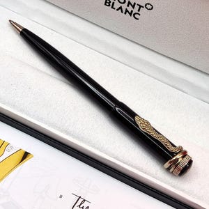 Pode incluir: Caneta Montblanc preta com design de cobra dourada. A caneta está em uma caixa branca com o logotipo Montblanc. A caneta tem uma ponta dourada e um corpo preto. A caneta tem aproximadamente 14 cm de comprimento.