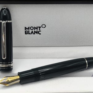 May include: A black Montblanc fountain pen with a gold and silver nib, featuring a matching black cap with a silver clip and band. The pen is displayed in a white presentation box with the Montblanc logo and brand name printed in black.