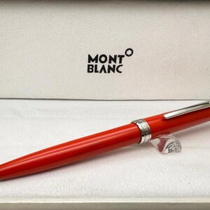 Puede incluir: Un bolígrafo de color rojo brillante con un clip plateado y una banda central plateada con detalles intrincados. El bolígrafo está colocado horizontalmente frente a una caja de presentación blanca que tiene el texto negro MONTBLANC impreso.