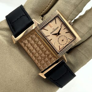 Puede incluir: Un reloj de pulsera rectangular de oro rosa con una esfera texturizada de color cobre, un subdial de segundos pequeños y el texto Jaeger-LeCoultre. El reloj cuenta con una correa de cuero negro y una caja giratoria con patrón geométrico.