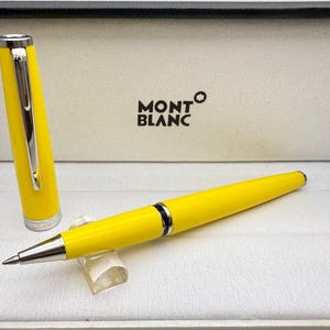 Op de afbeelding: Een felgele Montblanc rollerballpen met een zilverkleurige clip en afwerking. De pen wordt getoond met de dop eraf, rustend op een kleine doorzichtige standaard. Het Montblanc-logo is in het zwart gedrukt in de open presentatiedoos.