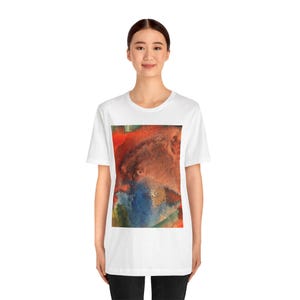 May include: A white short-sleeved t-shirt featuring a rectangular graphic print on the chest. The graphic displays an abstract composition with blended washes of vibrant red, deep orange, dark blue, and hints of green in a textured style.