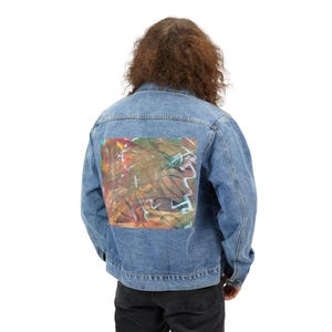 May include: A light blue denim jacket with a colorful abstract art design on the back. The artwork features a mix of colors including red, green, and yellow. The jacket has a classic collar and button-up front.