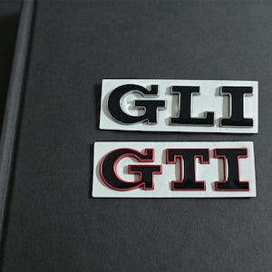 Peut inclure: Deux emblèmes adhésifs rectangulaires avec des lettres en gras. L'emblème supérieur affiche les lettres GLI en noir avec des contours argentés. L'emblème inférieur affiche les lettres GTI en noir avec des contours rouges sur fond blanc.