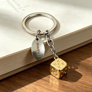 Op de afbeelding: Een zilverkleurige metalen sleutelhanger met een ronde ring, een kleine ovale bedel gegraveerd met de naam "merrick" in kleine letters, en een goudkleurige zeszijdige dobbelsteenbedel aan een korte ketting, alles met een gepolijste afwerking.