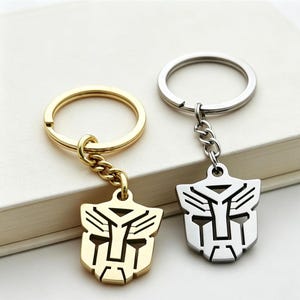 Peut inclure: Deux porte-clés en métal représentant le logo Autobot de Transformers. L'un est de couleur dorée avec un anneau et une chaîne assortis, tandis que l'autre est de couleur argentée avec un anneau et une chaîne assortis. Les pendentifs sont plats.