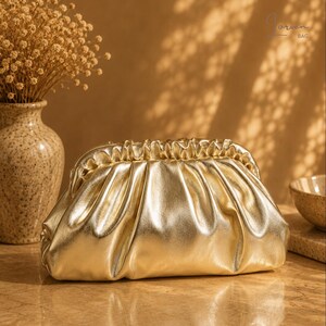 Può includere: Una pochette color oro metallizzato con chiusura superiore arricciata e una silhouette morbida e rilassata. La borsa presenta una superficie liscia e riflettente con un'apertura a telaio. Il testo "Lorven BAG" è visibile in alto a destra.