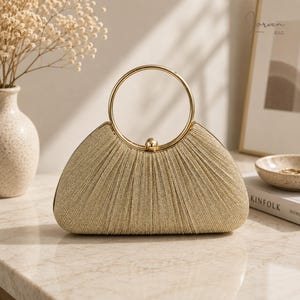Può includere: Una pochette color oro con una texture in tessuto plissettato scintillante e un manico circolare in metallo. La borsa presenta una silhouette semicircolare e una piccola chiusura sferica in metallo al centro superiore per la chiusura.