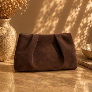 May include: A dark brown suede clutch bag with a soft, pleated front design. The rectangular bag features a smooth, matte texture and a structured frame, showcasing a clean, minimalist aesthetic without any visible hardware or external closures.