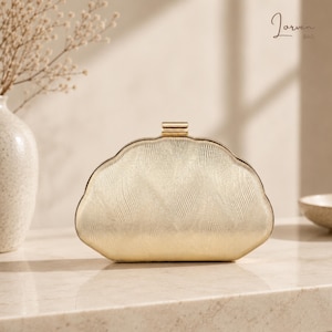 May include: A gold-colored clutch bag with a curved, shell-like silhouette and a textured, wavy surface pattern. The bag features a metallic gold frame and a rectangular clasp closure at the top center. The brand name Jorven Bag is visible.