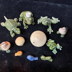 May include: A collection of small ceramic figurines, including a green turtle, frogs, birds, and a small orange fish. The figurines are in various poses and colors, with a neutral-colored sphere in the center.