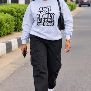 May include: A light gray crewneck sweatshirt featuring the black text AINT NO FAMILY LIKE THE ONE I GOT, paired with black cargo pants, white sneakers, a black shoulder bag, and black sunglasses. The sweatshirt has long sleeves and a ribbed hem.