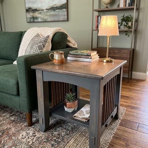 May include: A wooden side table with a dark gray base and a natural wood top. The table has a lower shelf and vertical slats on the sides. A lamp, books, and a mug sit on top. The table is 24 inches wide, 24 inches deep, and 24 inches high.