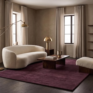 May include: A curved cream-coloured sofa with textured upholstery, a matching cream-coloured armchair with a dark wood base, a rectangular dark wood coffee table, a brass floor lamp, and a brass table lamp, all arranged on a large deep purple rug.