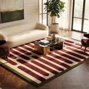 May include: A rectangular area rug featuring a repeating pattern of dark red rounded horizontal bars on a cream background, framed by a thin dark green border. The rug is positioned on a dark wood floor beneath a coffee table and seating.