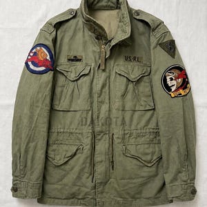 May include: An olive green field jacket featuring a high collar, a front zipper, and four button-flap pockets. The jacket is adorned with various patches, including a winged creature, a skull with goggles, and the text U.S.-R.L. on the chest.