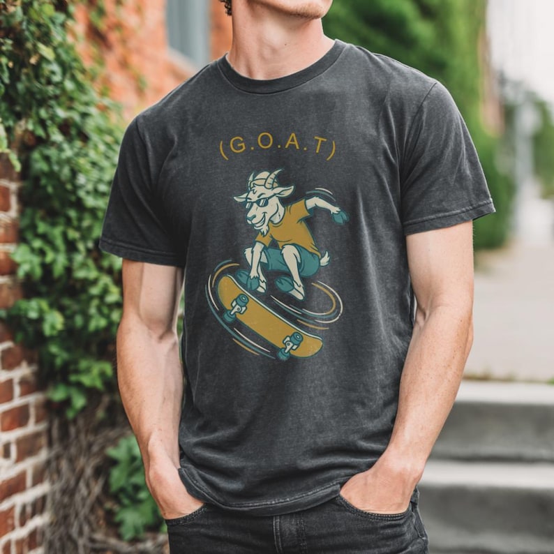 Może przedstawiać: Ciemnoszary t-shirt z kr&oacute;tkim rękawem z grafiką przedstawiającą kozę w okularach przeciwsłonecznych jadącą na deskorolce. Nad kozą znajduje się napis G.O.A.T. w złotym kolorze. Koza ma na sobie musztardową koszulkę i turkusowe szorty.