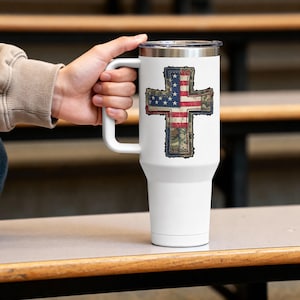 Könnte beinhalten: Ein weißer Edelstahlbecher mit Henkel und durchsichtigem Deckel, der in der Mitte mit einem Kreuzmotiv bedruckt ist. Das Kreuzdesign kombiniert ein rot-weiß-blaues amerikanisches Flaggenmuster mit grünen Camouflage-Elementen im unteren Bereich.