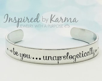 May include: Silver cuff bracelet with the inscription "be you... unapologetically" engraved on it. The bracelet is on a light blue background.
