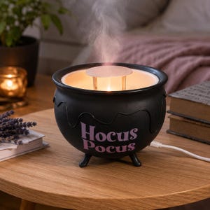 Könnte beinhalten: Ein schwarzer, kesselartiger elektrischer Duftdiffusor mit drei kleinen Standfüßen und einem tropfenförmigen Rand. Auf der Vorderseite steht der violette Schriftzug Hocus Pocus. Ein weißer Nebel steigt aus der Mitte auf, die ein Licht enthält.