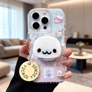 May include: Clear phone case with a cute cartoon seal design. The case features a white seal with a black smiling face, pink cheeks, and a pop-out grip. The case has a light blue and pink background with cartoon characters.