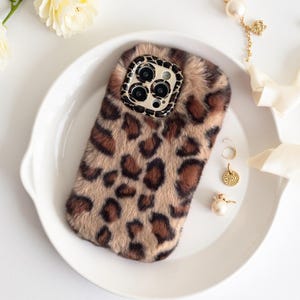 May include: A rectangular smartphone case covered in a soft, faux fur material featuring a brown and black leopard print pattern. The camera cutout area is framed by a matching leopard print border, surrounding the three circular camera lenses.