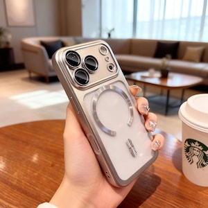 Op de afbeelding: Een zilverkleurige smartphonehoes met een metalen frame, een transparant achterpaneel en een cirkelvormige magnetische oplaadring. Het cameramodulegedeelte is versterkt met een metalen rand en onder de ring zit een decoratief accent.