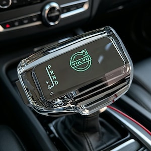 May include: A clear crystal gear shifter knob for a Volvo vehicle featuring an illuminated green Volvo logo and gear selection indicators including P, R, N, D, and S with plus and minus symbols. The shifter has a polished, transparent finish.