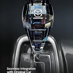 May include: A clear, faceted crystal gear shift knob for a Mazda vehicle, featuring the Mazda logo embedded within the top. The knob is positioned above a gear selector console marked with the letter P. Text on the image reads: Seamless Integration with Original Car.