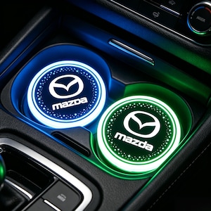 Puede incluir: Dos insertos iluminados para portavasos de coche con el logotipo de Mazda. Uno brilla en azul, el otro en verde. El logotipo y el texto de Mazda son blancos. Los insertos están rodeados por un anillo de pequeñas estrellas blancas.