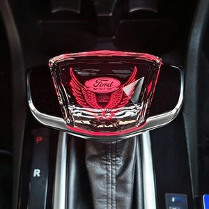 May include: A clear, rectangular gear shift knob featuring a red illuminated Ford logo with stylized wings. The knob has a polished black base and is mounted on a black leather-textured gear shift boot within a vehicle center console.
