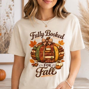 May include: A cream-coloured short-sleeve t-shirt featuring a graphic of a pumpkin constructed from stacked books. The text "Fully Booked FOR Fall" is printed in brown script above and below the pumpkin, which is accented with autumn leaves.