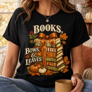 Pode incluir: Uma t-shirt preta com uma estampa gráfica de uma pilha de livros amarrados com um grande laço laranja, rodeada por folhas de outono e abóboras. O texto "BOOKS, BOWS, & LEAVES" está impresso com uma fonte decorativa sobre os livros.