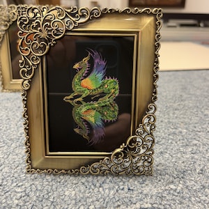 May include: A gold-toned metal picture frame with an ornate design. The frame holds a black background with a colorful image of a green dragon with rainbow wings.