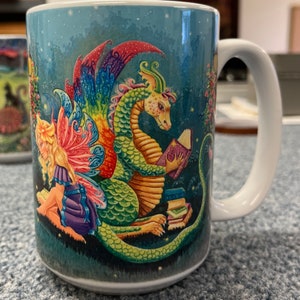 May include: A white ceramic mug with a colorful illustration of a fairy and a dragon reading books. The background is a dark blue with stars and a green forest.