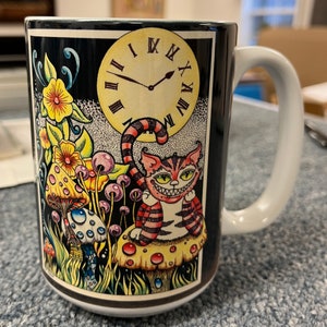 May include: White ceramic mug with a black handle. The mug features a colorful illustration of a Cheshire Cat with a mischievous grin, surrounded by mushrooms and a clock face with Roman numerals. The background is black.