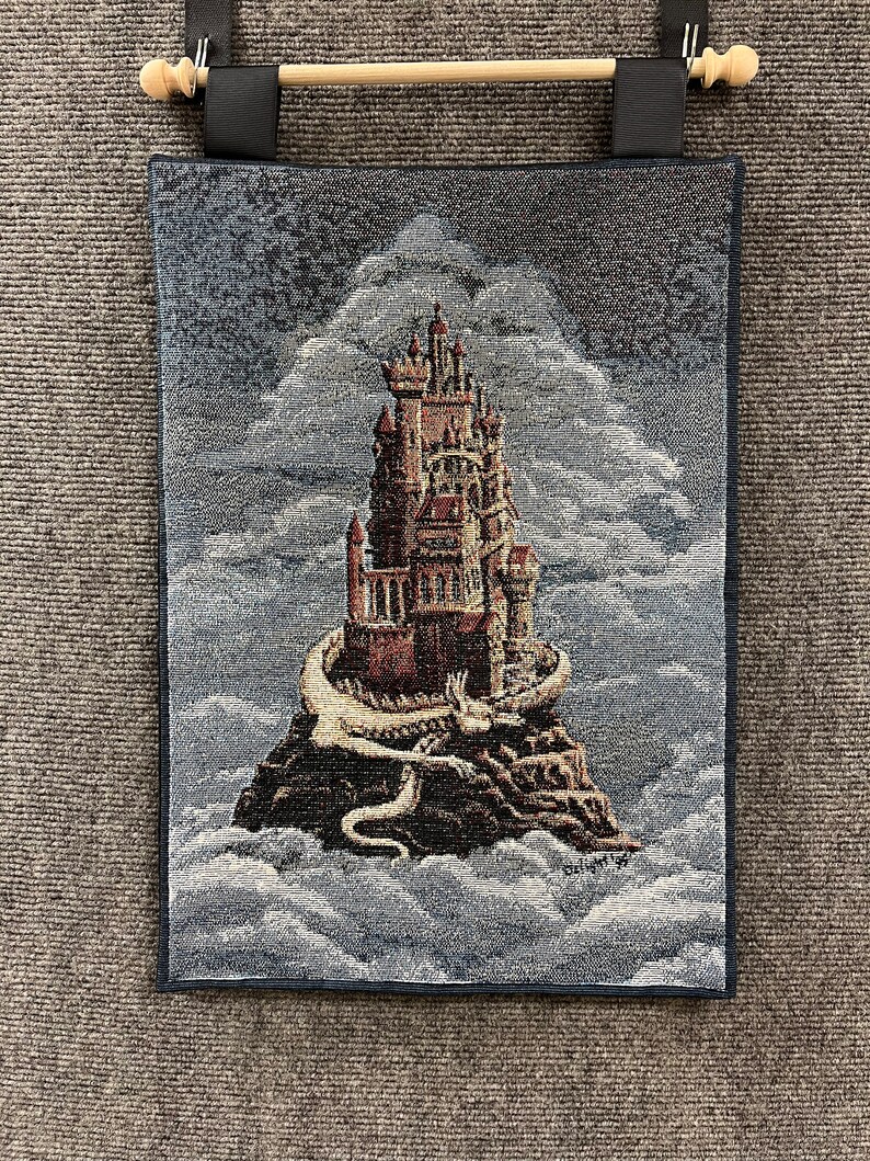 Puede incluir: Tapiz de pared con un castillo de fantas&iacute;a sobre una isla rocosa rodeada de nubes. El castillo es marr&oacute;n y rojo con detalles intrincados. Un drag&oacute;n blanco envuelve la isla. El tapiz tiene un borde azul oscuro y una barra de madera para colgar.