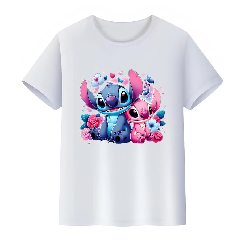 K&ouml;nnte beinhalten: Ein wei&szlig;es kurz&auml;rmeliges T-Shirt mit einem zentralen Grafikdruck von zwei Zeichentrickfiguren, einer blauen und einer rosa, die zusammen sitzen, umgeben von bunten Blumen, kleinen Herzen und Schmetterlingen in Blau- und Rosat&ouml;nen.