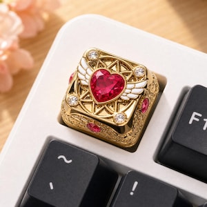 Peut inclure: Une touche de clavier mécanique dorée ornée d'une pierre précieuse en forme de cœur rouge au centre, flanquée d'ailes blanches. La touche carrée présente un travail de filigrane métallique, des cristaux clairs aux coins et des gemmes rouges.
