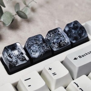 May include: Four custom mechanical keyboard keycaps featuring a black base with a translucent, swirling white smoke or marble-like resin top. These artisan keycaps are positioned in a row on a white keyboard next to a standard Backspace key.