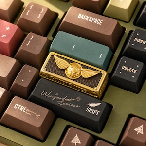 May include: A mechanical keyboard with custom keycaps in shades of brown, green, and black. Features include a gold-coloured Golden Snitch keycap, a black Shift key with the text Wingardium Leviosa and a feather icon, and a Ctrl key labelled Unicorn hair.